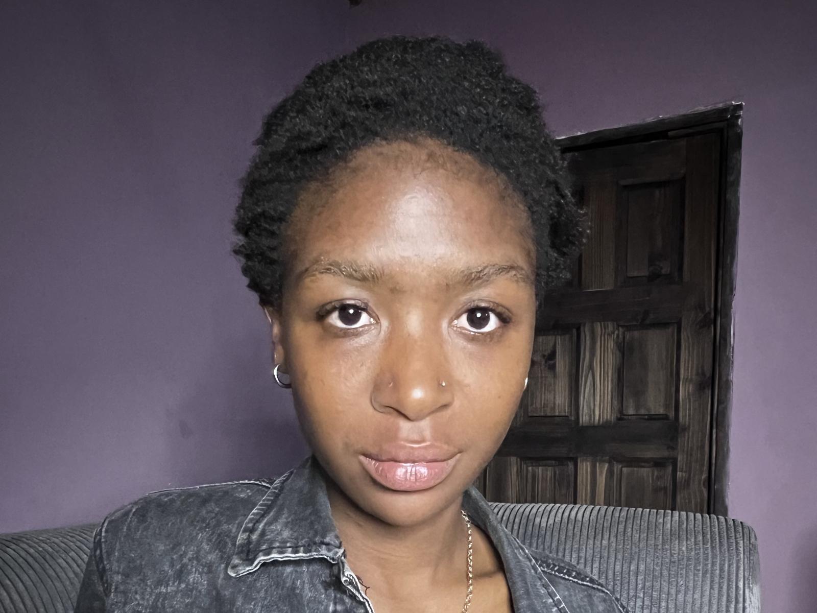 CupcakeDolly has a dark brown complexion, short curly black hair, small hoop earrings, and is wearing a slightly unbuttoned denim jacket with pinkish lips.