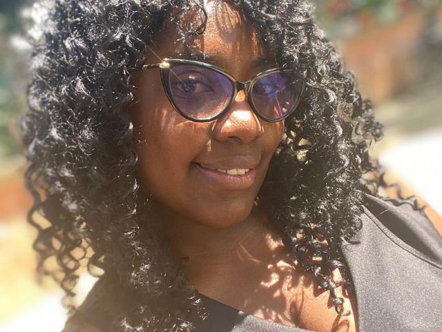 CynthiaRamos with dark curly hair, wearing black sunglasses and a black top, smiling in sunlight. Her brown skin and small, white teeth are visible.