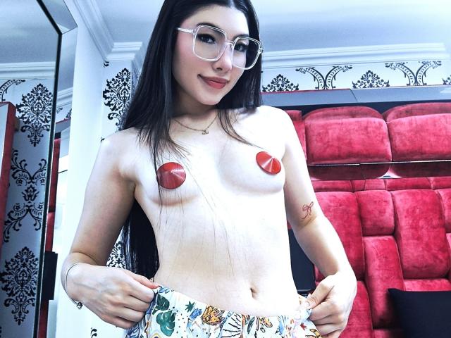 ViolettaLeroy, topless with red nipple pasties, wearing glasses, has long black hair, small tits, and a floral skirt, standing in a room with red velvet chairs.