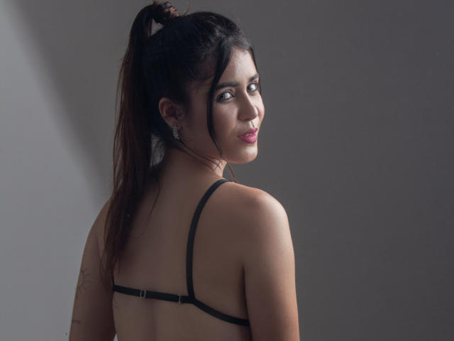 EvaSingh with brown ponytail, black bra straps, earrings, looking over shoulder, pink lipstick, light skin, small tits, subtle smile, gray background, soft light, photograph.