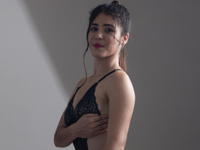 EvaSingh with dark hair in a ponytail, wearing a black lace bra, covering her small tits with one hand, smiling, and looking to the side.