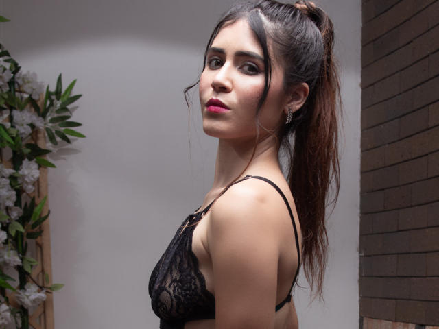 EvaSingh with dark brown hair in a ponytail, wearing black lace bra, showing her tits, looking over shoulder, red lipstick, small earrings, standing next to a floral arrangement.