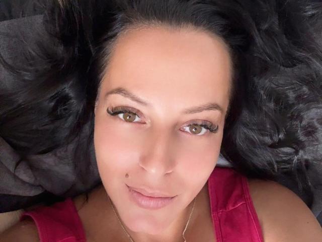 ShawtLana with black hair, brown eyes, and a small beauty mark on her left cheek, wearing a pink top, lying down, looking at the camera with a slight smile.