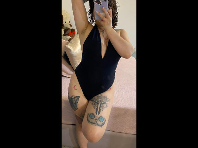A selfie of AnikaFleur in a black, deep-v, one-piece swimsuit. She has wavy, brown hair and is holding a smartphone in front of her face. Her tattoos include a crescent moon on her right thigh, a tree with eyes on her left thigh, and a butterfly on her left calf. She stands in a room with a pink quilted bed in the background and a stuffed bear on the left. The image is framed with black borders. Her skin is light. The focus is on her tattoos and swimwear.
