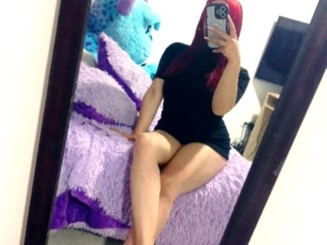 AshleyPlatta with red hair, black short dress, and bare legs, sitting on a purple fuzzy bed, taking a mirror selfie, showing her ass and pussy slightly visible.