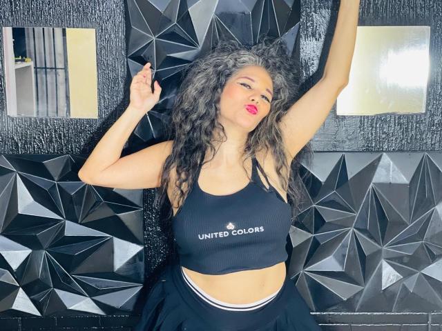 CuteMatureSassy with curly gray hair, red lipstick, black sports bra, and black pants, arms raised, standing against geometric black wall, showing her toned midsection and small tits.