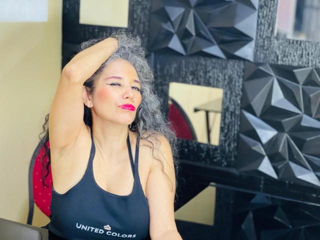 CuteMatureSassy with gray curly hair, black sports bra, pink lipstick, and raised arm, showing off her tits and confident smile. Black geometric wall in background.