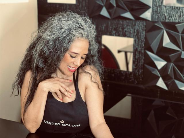 CuteMatureSassy with gray curly hair, smiling, wearing a black United Colors sports top, touching her chest, red nails, sitting, black geometric-patterned background.