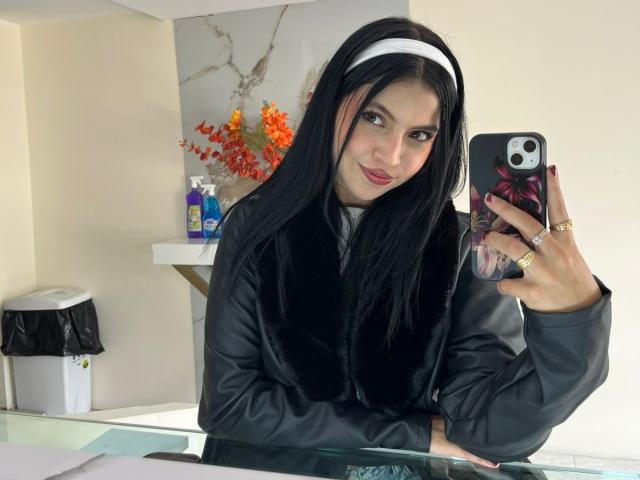 NyraVanel, con cabello negro largo, diadema blanca, chal de terciopelo negro, sosteniendo un teléfono, tomando una selfie, sonriendo, chaqueta gris, anillos dorados, esmalte de uñas rojo, en un baño moderno..