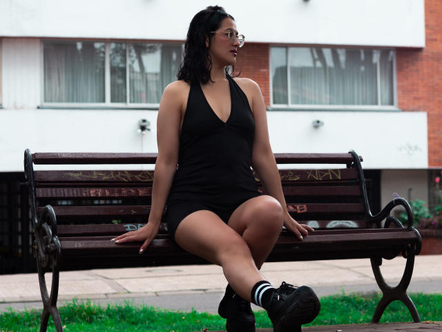 ElizabethDupont sits on a wooden bench wearing a black halter dress, glasses, and black sneakers with white socks. She has medium tits, black curly hair, and a curvy ass.