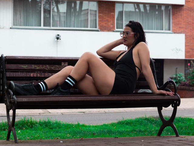 ElizabethDupont with dark skin and glasses, wearing a black one-piece and black boots with blue stripes, lounging on a bench, her ass and tits visible, hand on her head.