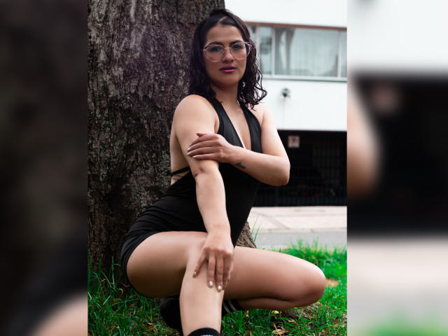 ElizabethDupont with dark wavy hair, glasses, black sleeveless dress, squatting by a tree, showing her ass, holding her arm, tattoo on her wrist, and a camera in the foreground.