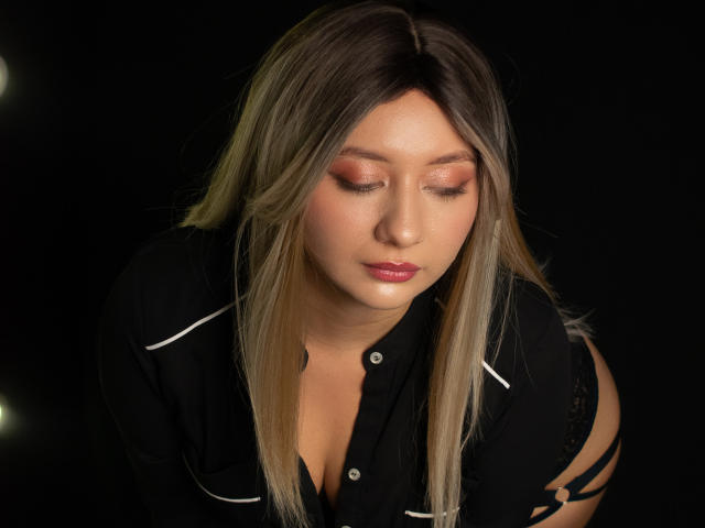 CataleyaBlow with blonde hair, closed eyes, and pink lipstick, wearing a black shirt with white trim, bending forward, showing a hint of black straps on her right side.