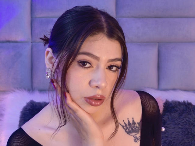 LindaMancini has dark hair in a messy bun, wearing a black top, with her hand on her cheek, showing a crown tattoo on her shoulder, and shiny lips.