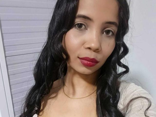 AngelicaHott has long black wavy hair, light brown skin, red lipstick, and a gold necklace, with a subtle smile and brown eyes. She's wearing a white top.