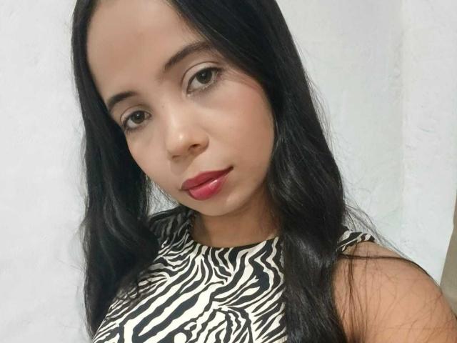 AngelicaHott has dark, wavy hair, brown eyes, and red lipstick. She's wearing a zebra-patterned top, and her smooth, tan skin is visible. Her tits are not shown.