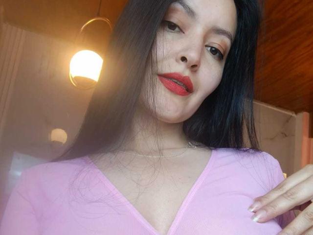 Aria, con lunghi capelli neri, una maglietta rosa, rossetto rosso, un leggero sorriso, una mano sul petto, che guarda la fotocamera, luce soffusa proveniente da una lampada a sospensione, soffitto in legno, selfie casual..