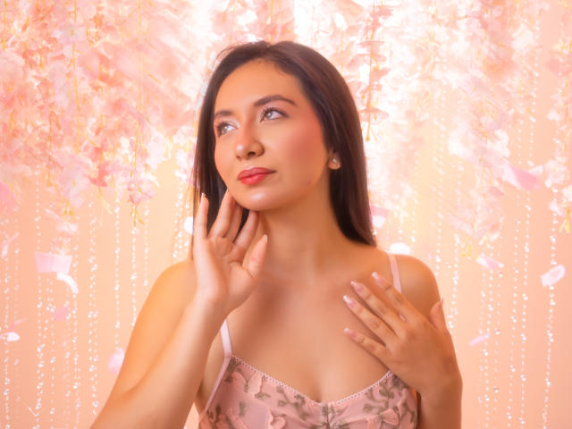 CataleyaBlow with long dark hair, pink floral dress, and red lipstick, touching her cheek with one hand, looking up thoughtfully. Her tits are small, her pussy is not visible.