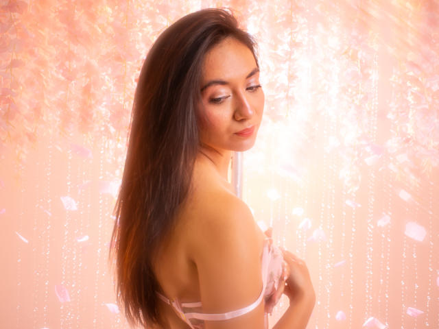 CataleyaBlow with long brown hair, pink eyes, and a gentle smile, wearing a pink strap, covering her small tits with her hand, against a soft pink, sparkling background.
