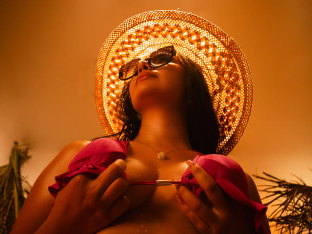 CataleyaBlow with dark skin, wearing a pink bra, holding it up to show her tits, brown wavy hair, and a woven hat, looking up with sunglasses on.