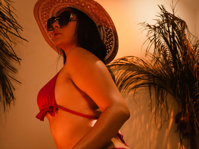 CataleyaBlow in a red bikini, wearing a straw hat and dark sunglasses, with her tits showing and a palm tree in the background, under warm orange lighting.