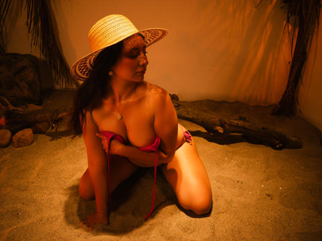 CataleyaBlow kneeling on sand, wearing a straw hat and pink bikini top, covering her big, round tits with her hands, looking to the side, under warm, orange lighting.