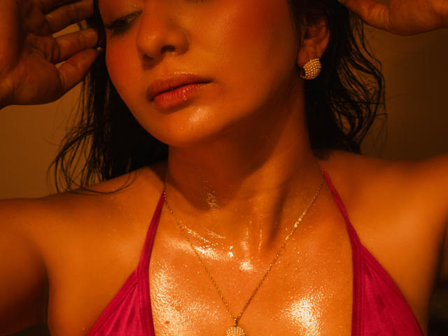 CataleyaBlow's glistening, shiny tits in a pink bikini top, wet skin, hands in hair, orange light, small diamond earrings, and a gold necklace with a heart pendant.