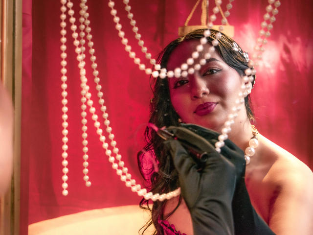 CataleyaBlow with wet dark hair, purple lipstick, black gloves, holding pearl necklace, smirking, red background, pearls in front, looking sexy, showing off tits, elegant, seductive.