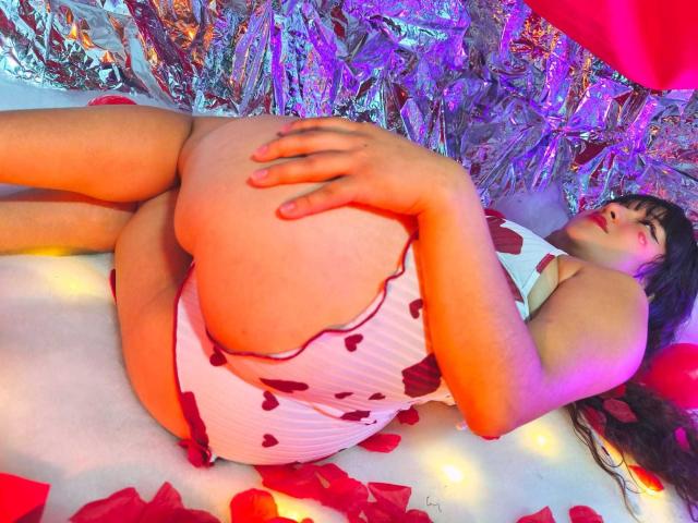 NicoleMartins lying on her side, wearing a white heart-patterned dress, gripping her big ass with one hand, surrounded by red rose petals and yellow fairy lights.