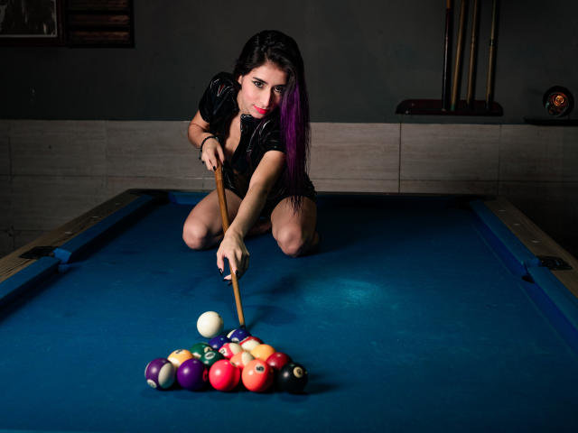 JuliaJobs kneels on a blue pool table, aiming a cue stick at a clustered ball set. She has long dark hair with purple streaks, wearing a black, shiny top. Her red lipstick contrasts with her dark makeup. The balls are colorful, with a mix of solids and stripes. The cue stick is wooden, and the table's blue felt is smooth. The background is dimly lit, with a wall and pool sticks in the back. The image is sharply focused on JuliaJobs and the balls.