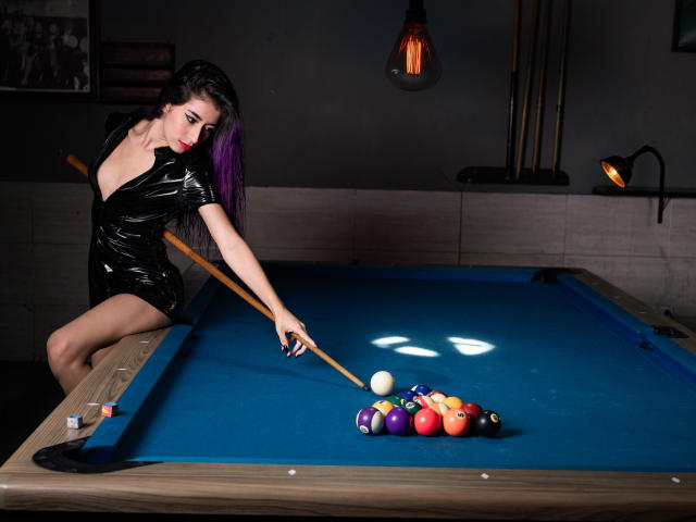 JuliaJobs stands by a blue-clothed pool table, holding a wooden cue stick and aiming at a ball cluster. She's wearing a shiny black vinyl dress with a deep V-neck, revealing her cleavage. Her long purple hair contrasts with her fair skin. A hanging light bulb casts a warm glow. The cue ball, eight-ball, and other colored balls are neatly arranged on the table. A black cue holder is on the right. The image is sharply focused, emphasizing the pool table's texture and JuliaJobs' confident pose.