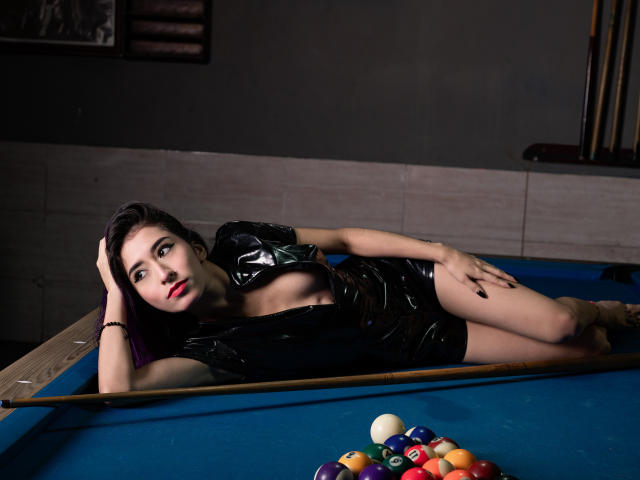 JuliaJobs, with black shiny dress and red lips, lies on blue pool table, legs crossed, hand on head, looking at camera, pool balls in front, dark room.