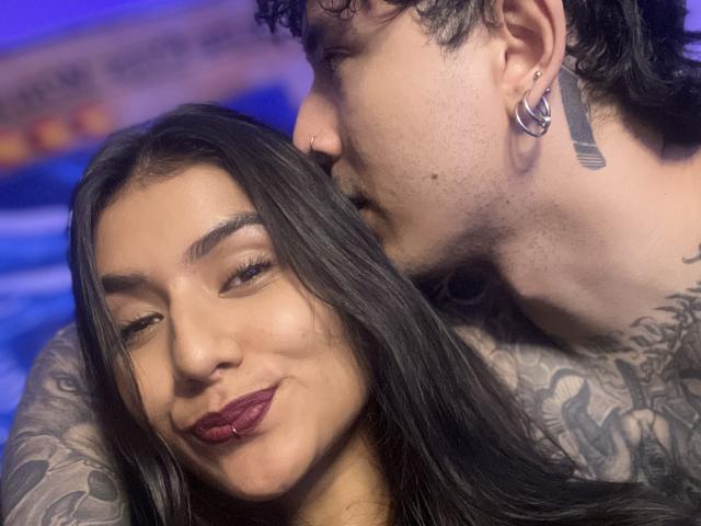 TheJoestars with long black hair and red lipstick, smiling, has a nose ring and tattooed arms, being gently kissed by TheJoestars with curly black hair and tattoos.