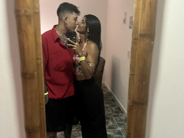 A mirror selfie of TheJoestars. A young man with short black hair and a red shirt kisses a young woman with long black hair and a black dress. She holds a phone with a floral case in her right hand, wearing pink scrunchies and a yellow wristband. The man has a silver chain necklace. The image is framed by wooden mirror edges. The floor is tiled in black and white patterns.