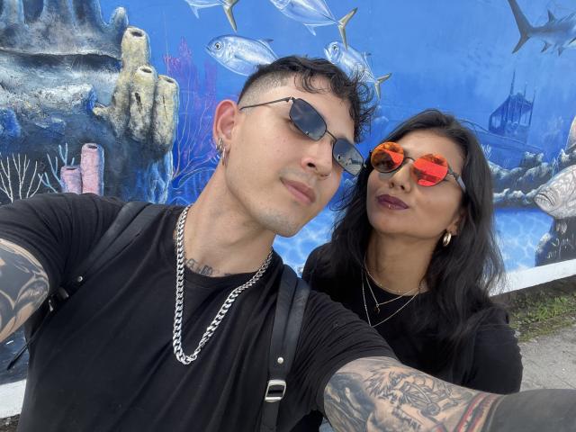 TheJoestars, a tattooed man with short black hair and sunglasses, stands next to a woman with long black hair and red-tinted glasses. Both wear black shirts and TheJoestars has a chain necklace. TheJoestars has a muscular build, and the woman has a medium build with dark lipstick. They stand in front of a vibrant blue mural with underwater fish and coral. TheJoestars has a confident expression, while the woman looks slightly surprised. The image is a selfie.