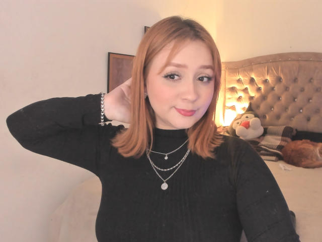 DarkCourtney met bruin haar dat tot op de schouders reikt, draagt een zwarte coltrui, een zilveren ketting en armband, en poseert met haar hand achter haar hoofd, glimlachend, op een bed met een gestoffeerde hoofdeinde..