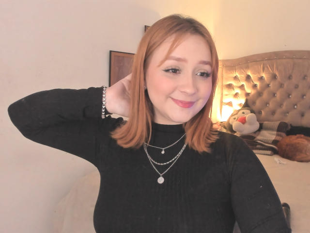 DarkCourtney, com cabelo ruivo na altura dos ombros, gola alta preta e vários colares, sorrindo, com a mão no cabelo, em um quarto aconchegante, um pássaro de pelúcia fofo na cama, luz suave..