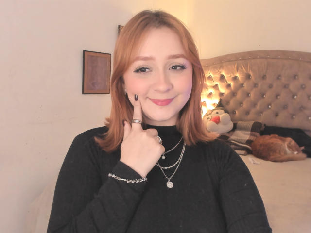 DarkCourtney has light red hair, black top, and is smiling with her finger on her cheek, showing a ring and necklace, with a cat and stuffed toy in the background.