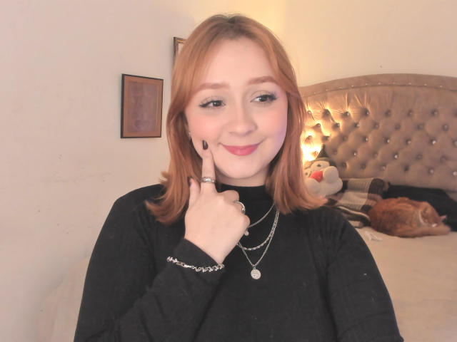DarkCourtney with red hair, black top, and jewelry, pointing at her cheek with a finger, smiling. She's got a cute face, and a sleepy orange cat on the bed.