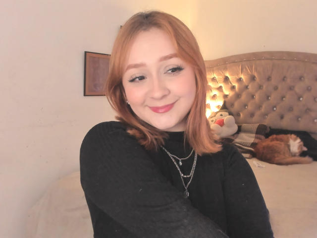 DarkCourtney with red hair, black top, and necklace, smiling at camera, bed with sleeping cat and stuffed animal in background, soft light, cute, attractive.