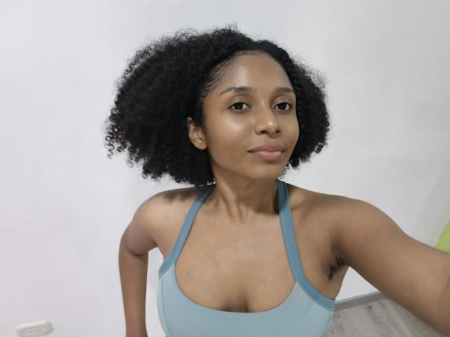 MeganWilliams with curly black hair, wearing a blue sports bra, showing her small boobs and armpit hair, in a selfie with a subtle smile.