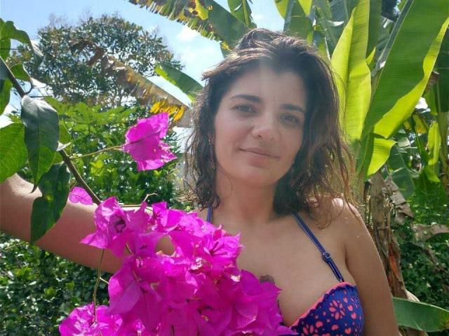 BulmaHot with dark, wavy hair, blue strap bikini, holding bright pink bougainvillea flowers, smiling subtly, standing in lush greenery, sunlight filtering through leaves.