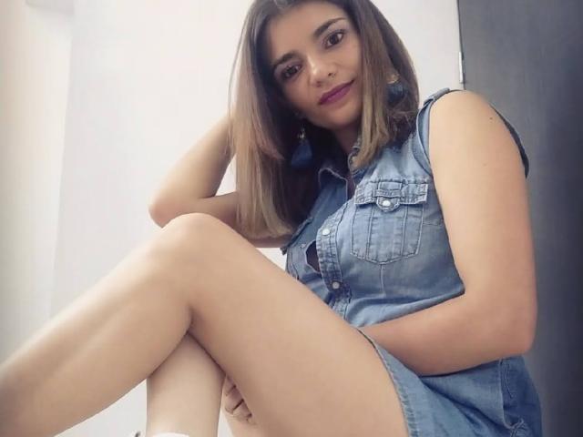BulmaHot with brown hair, wearing a denim sleeveless dress, sitting with legs crossed, showing her ass, dark purple lipstick, and blue earrings, smiling at the camera.