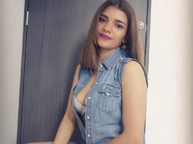 BulmaHot with medium tits, wearing a denim sleeveless vest, showing a hint of a white bra, brown hair, pink lipstick, and earrings, standing in front of a dark door.