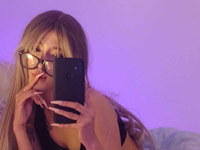 OliviaMonroee with long blonde hair, wearing glasses, black top, lying down, taking a mirror selfie, finger near her lips, purple lighting, holding a black phone, visible breasts.