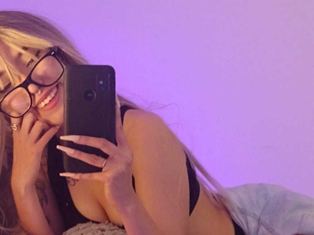 OliviaMonroee with glasses, smiling, holding a phone, showing her tits in a black top, lying on a bed, purple lighting, long blonde hair, tattoo on her arm.