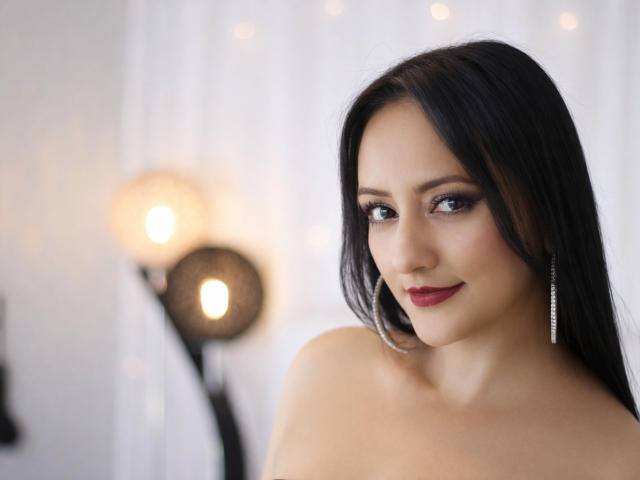 SarahTemptation with dark hair, red lipstick, and silver earrings, showing off her tits in a soft, white-lit room. She's smiling subtly, looking at the camera.