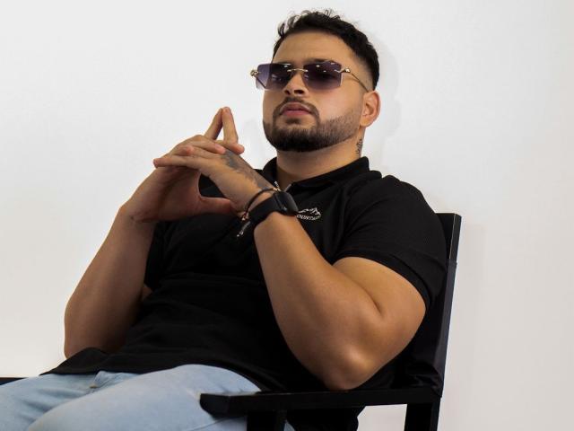 AryanBrown, avec une barbe, portant des lunettes de soleil, un t-shirt noir et un jean bleu, est assis sur une chaise noire, les mains jointes, regardant légèrement sur le côté, un tatouage au poignet visible..