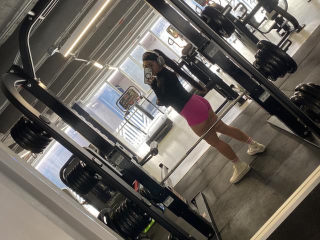 MiaDuboisX with toned ass, wearing pink booty shorts, white sneakers, black top, headphones, taking mirror selfie in gym, showing fit legs, reflecting weightlifting equipment.
