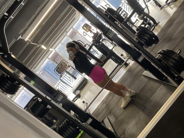 MiaDuboisX stands in a gym mirror selfie, showing her pink booty shorts, black top, and white sneakers, with a weight rack and barbells in the background.